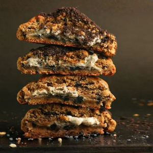 Four stacked triangular baked cookies or breads cut open to show a thick cream-colored filling with dark cookie pieces mixed throughout, set on a dark surface against a dark brown background.