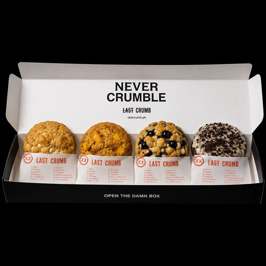 A black and white Last Crumble bakery box containing four assorted muffins or cookies, with the brand name 'NEVER CRUMBLE' printed on the inside lid.
